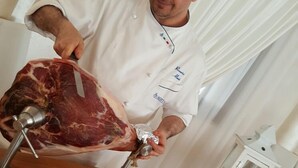 Un chef corta carne sobre una tabla con un plato de carne en rodajas y varios platos de fondo.