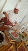 Un chef corta carne sobre una tabla con un plato de carne en rodajas y varios platos de fondo.