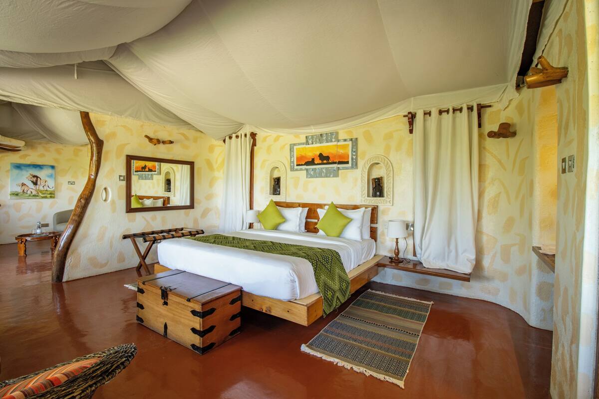 A spacious room with a large bed, wooden headboard, and a wooden chest of drawers. There is a small table and a chair in the corner, and a large window with curtains.