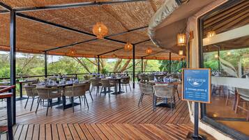 An outdoor dining area with tables and chairs, a thatched roof, and hanging lights.