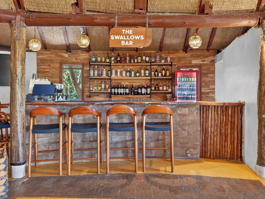 Zona de bar con taburetes de madera, barra y estanterías llenas de botellas.