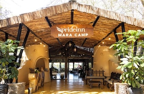 Eine rustikale Lodge mit einem strohgedeckten Dach, einem Schild mit der Aufschrift "prideinn Mara Camp" und einem gemütlichen Innenraum mit Sitzgelegenheiten und Pflanzen.