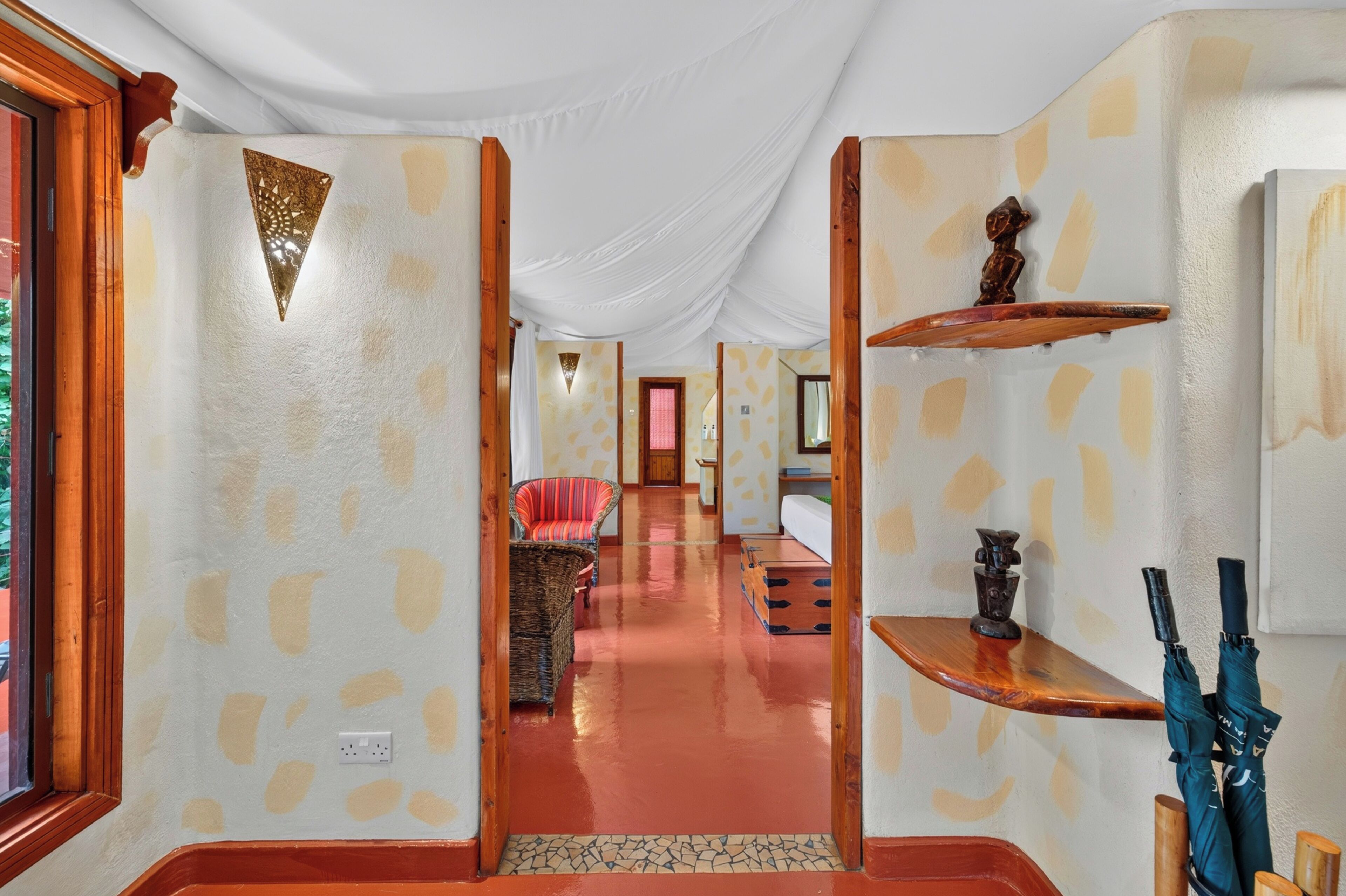 A corridor with a patterned wall, a red chair, a wooden shelf with an umbrella, and a small statue.