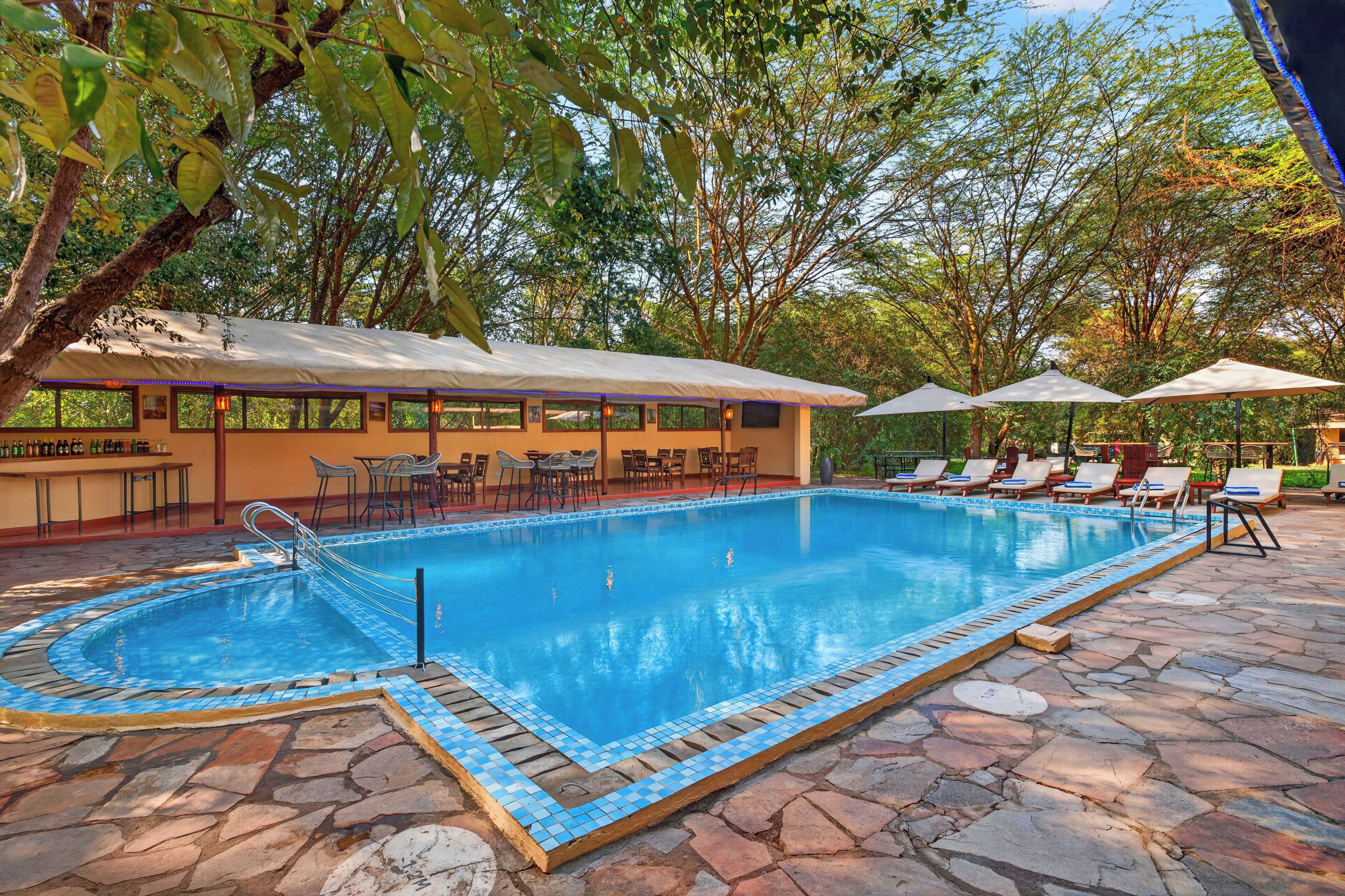 A swimming pool with a tiled edge, surrounded by lounge chairs and umbrellas, with a building in the background.