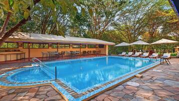 A swimming pool with a tiled edge, surrounded by lounge chairs and umbrellas, with a building in the background.
