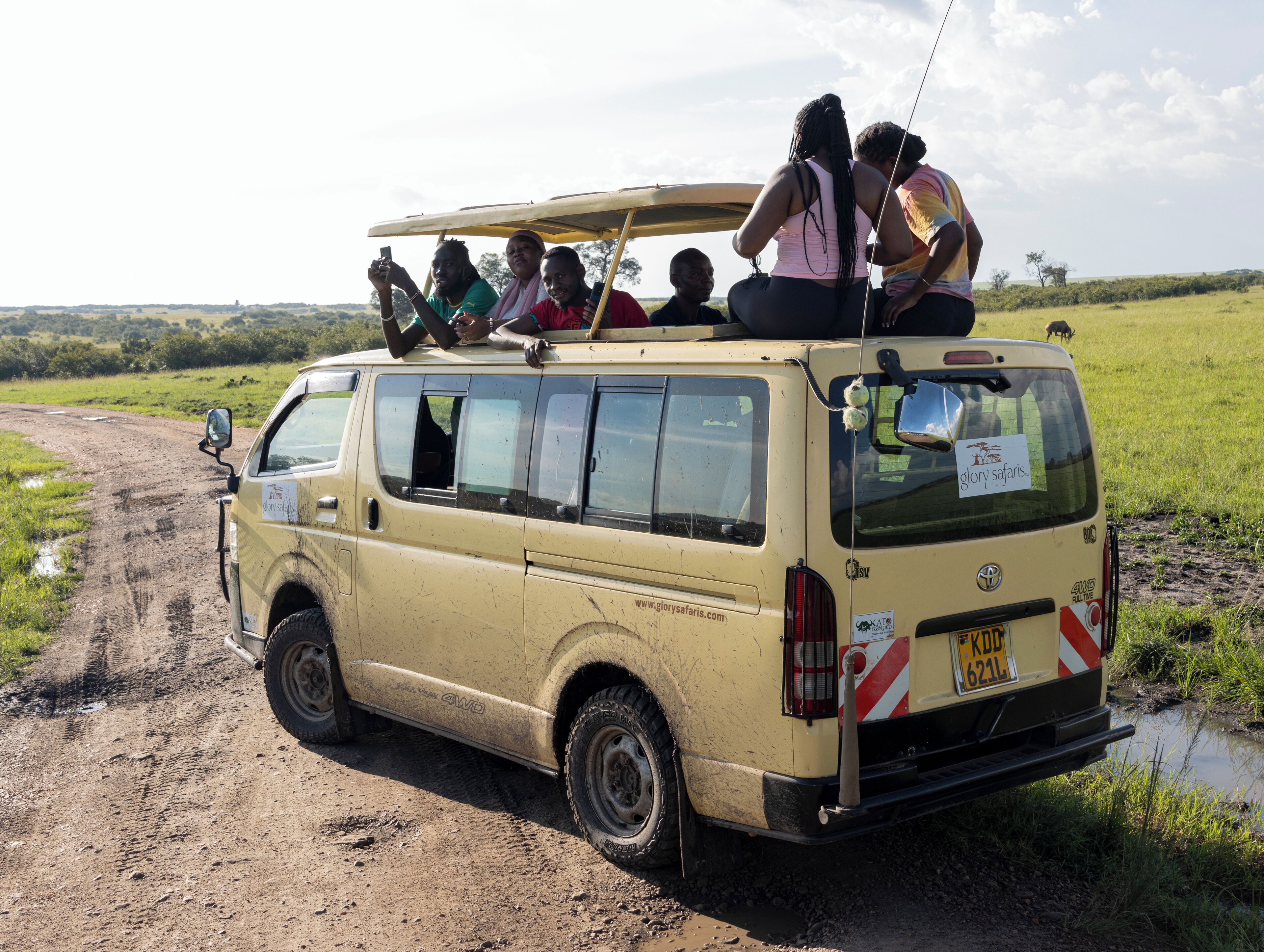 A beige van with a roof rack and passengers on top, parked on a muddy road with a grassy field and a small body of water in the background.