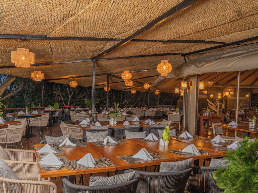 Ein Restaurant im Freien mit Holztischen und Korbstühlen unter einem reetgedeckten Dach mit hängenden Laternen.
