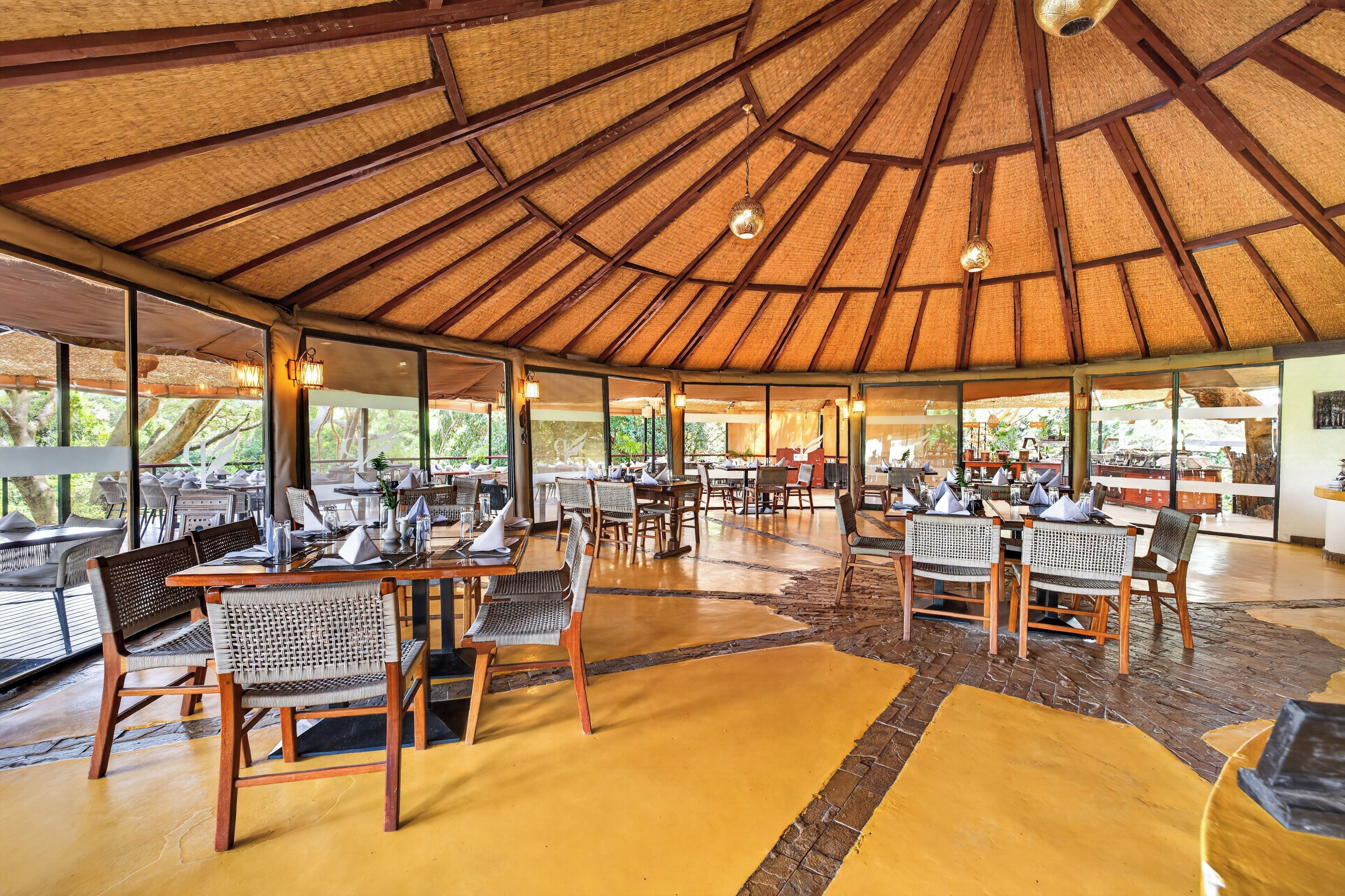 Un comedor interior con mesas y sillas, techo de paja y ventanas amplias.