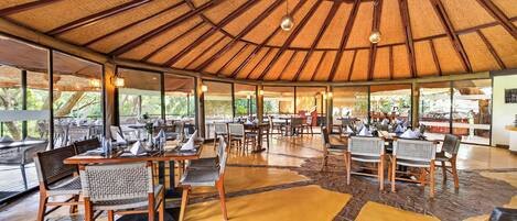Un comedor interior con mesas y sillas, techo de paja y ventanas amplias.