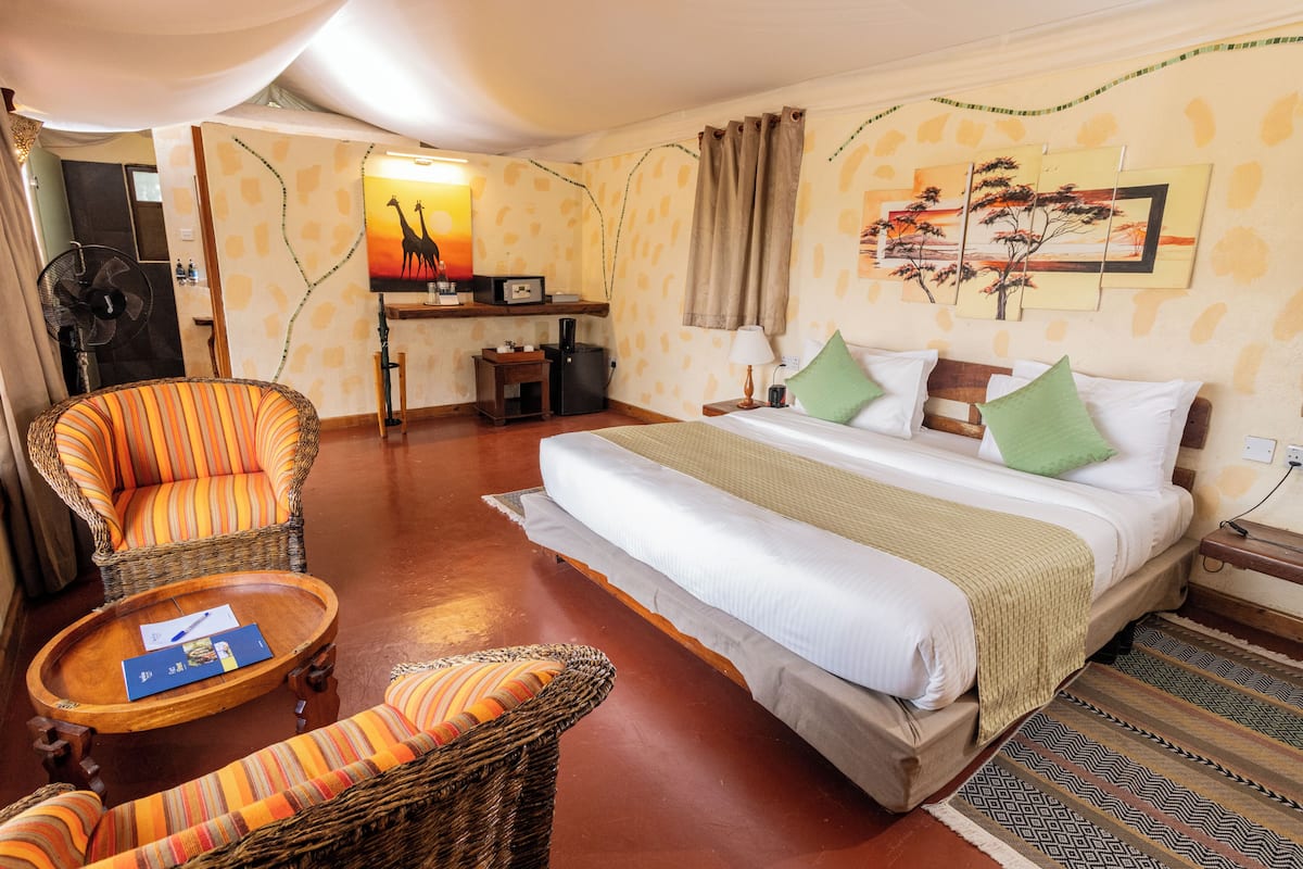 A hotel room with a bed, two wicker chairs, a small table, a desk with a fan, and artwork on the wall.