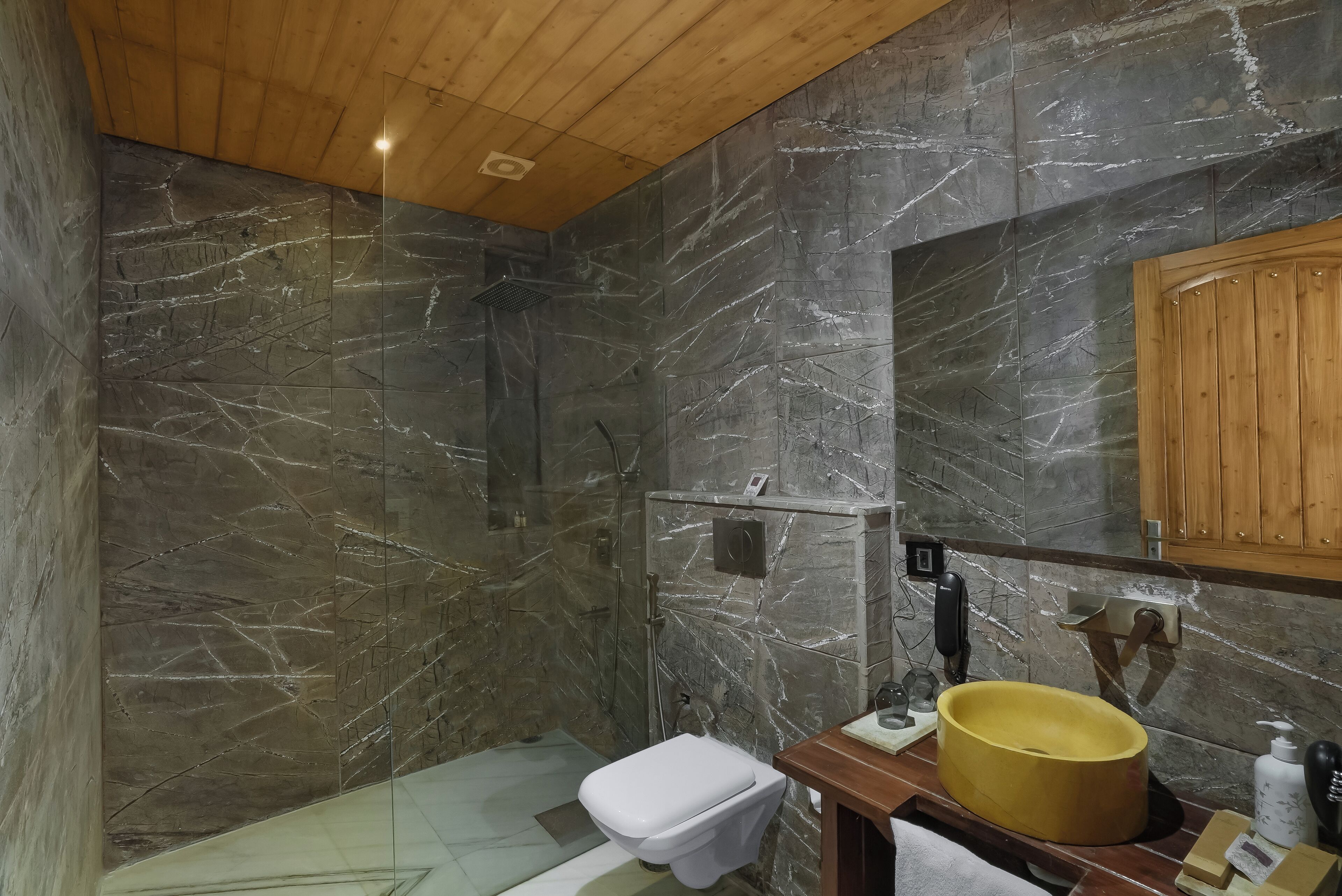 Baño con pared de ducha de mármol, inodoro y tocador de madera con lavabo amarillo.