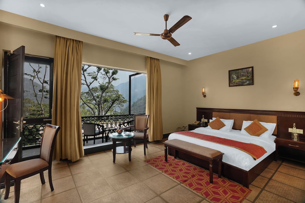 Quarto de hotel com uma cama grande, uma escrivaninha com cadeira, uma varanda com vista para árvores e montanhas, e um ventilador de teto.