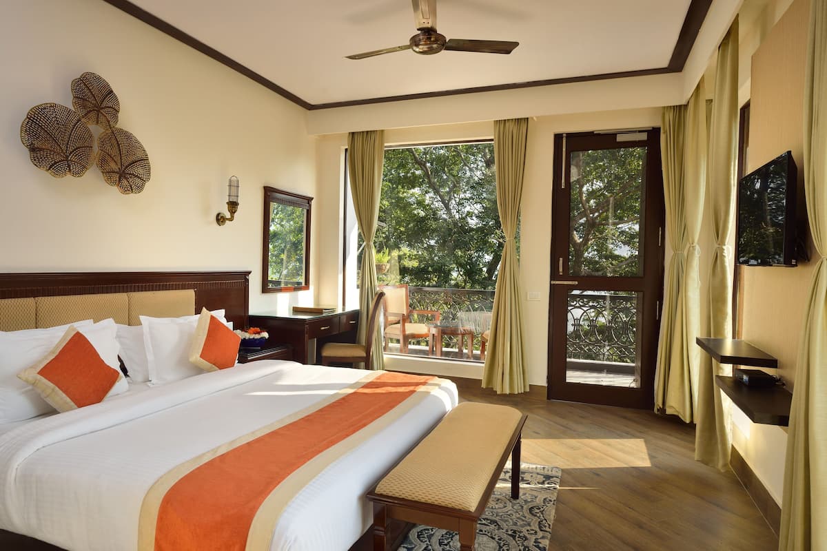 Quarto de hotel com uma cama grande, uma mesa, uma cadeira, uma TV e uma varanda com vista para árvores.