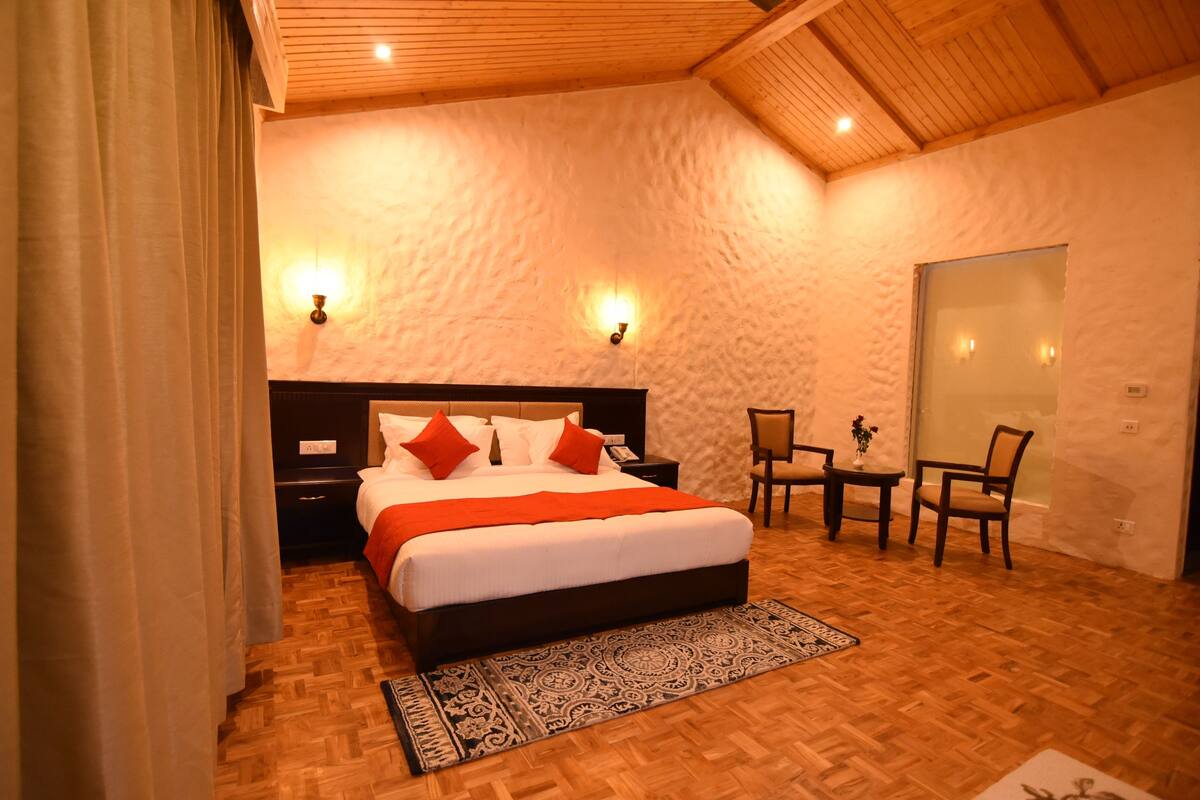 Quarto com teto de madeira, cama grande com roupa de cama vermelha e branca, duas luminárias de cabeceira, uma pequena mesa com cadeiras e um tapete estampado.