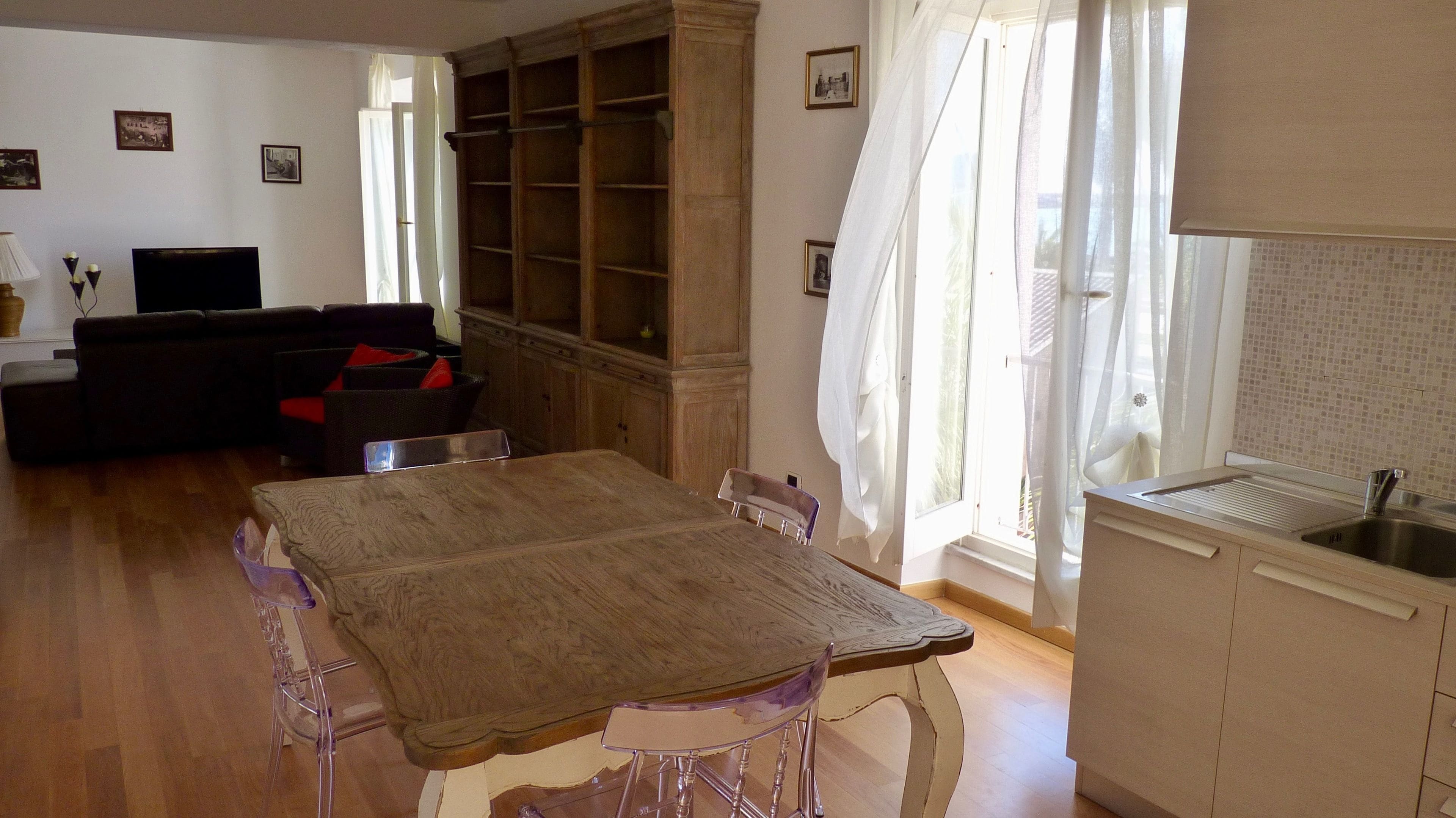Een eethoek met een houten tafel en stoelen, een keuken met een spoelbak, en een woonkamer met een bank en een televisie.
