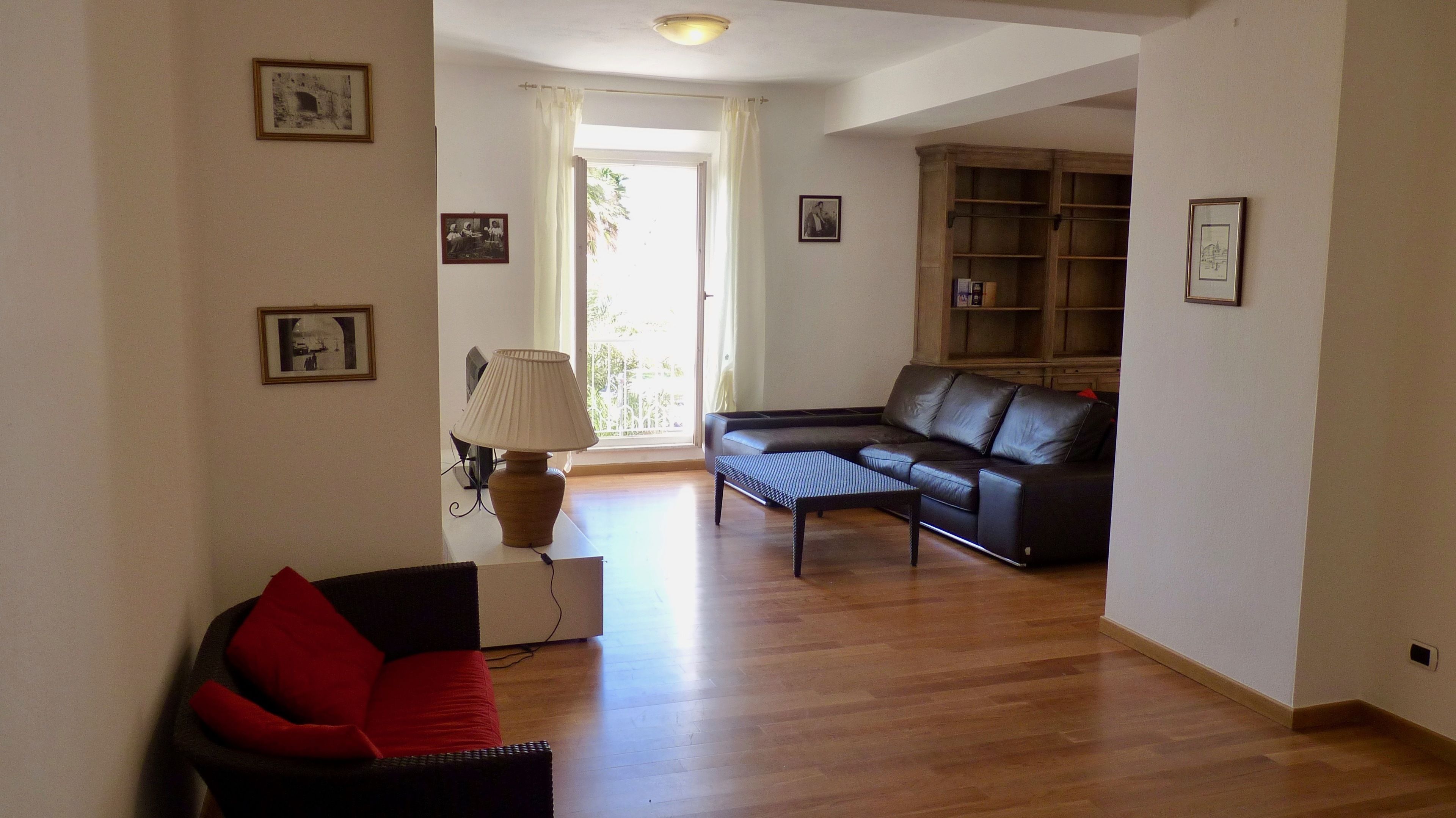 A living room with a black leather sofa, a red cushion, a wooden coffee table, and a lamp. There is a bookshelf and a door leading to a balcony.