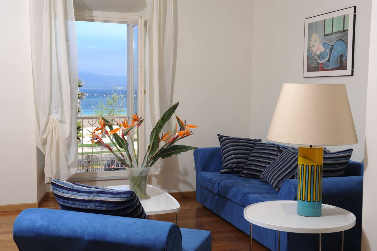 Una sala de estar con un sofá azul, una mesa de centro blanca, una lámpara y vista al mar a través de una ventana.