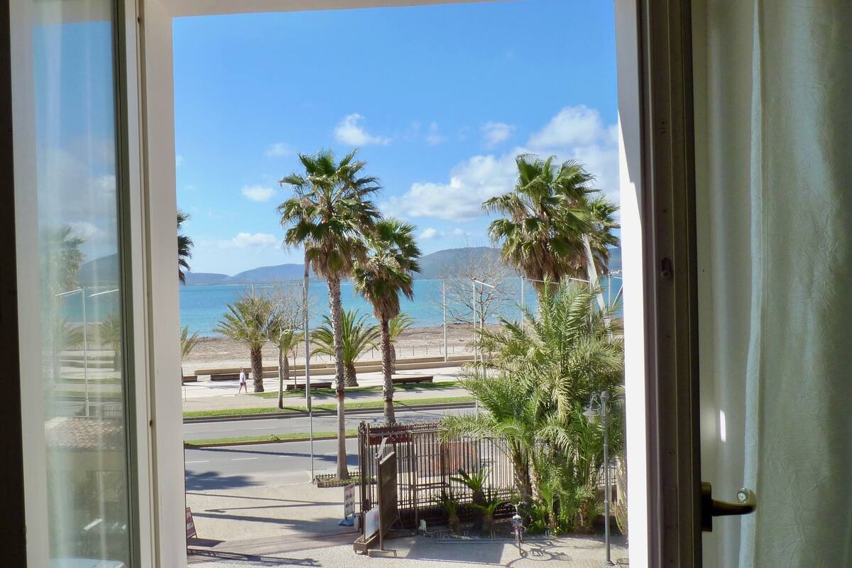 Vista a través de una puerta que da a un balcón con palmeras, una playa y un cielo despejado.