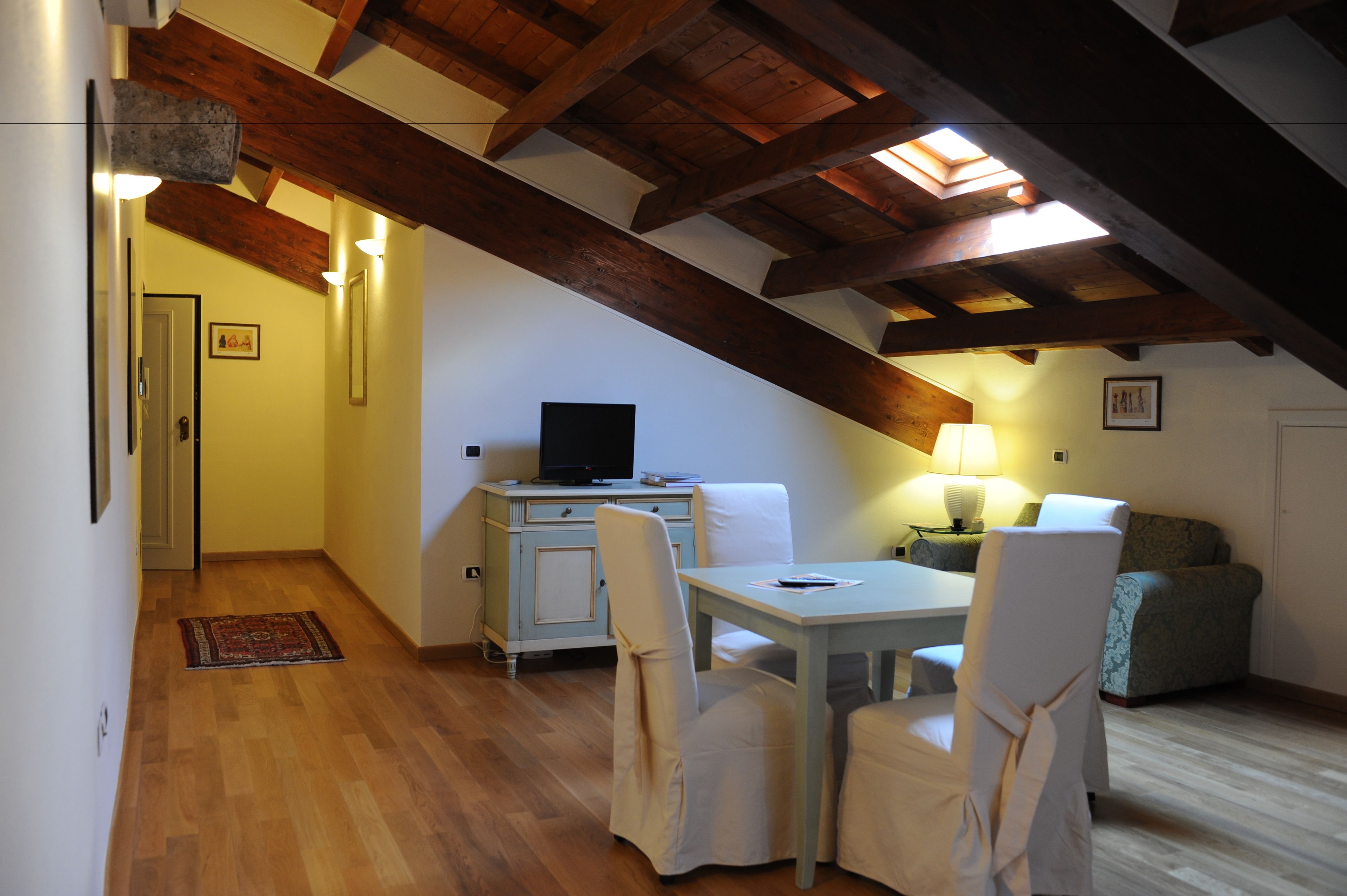 A cozy attic bedroom with a wooden ceiling, a desk, a chair, a sofa, and a TV.