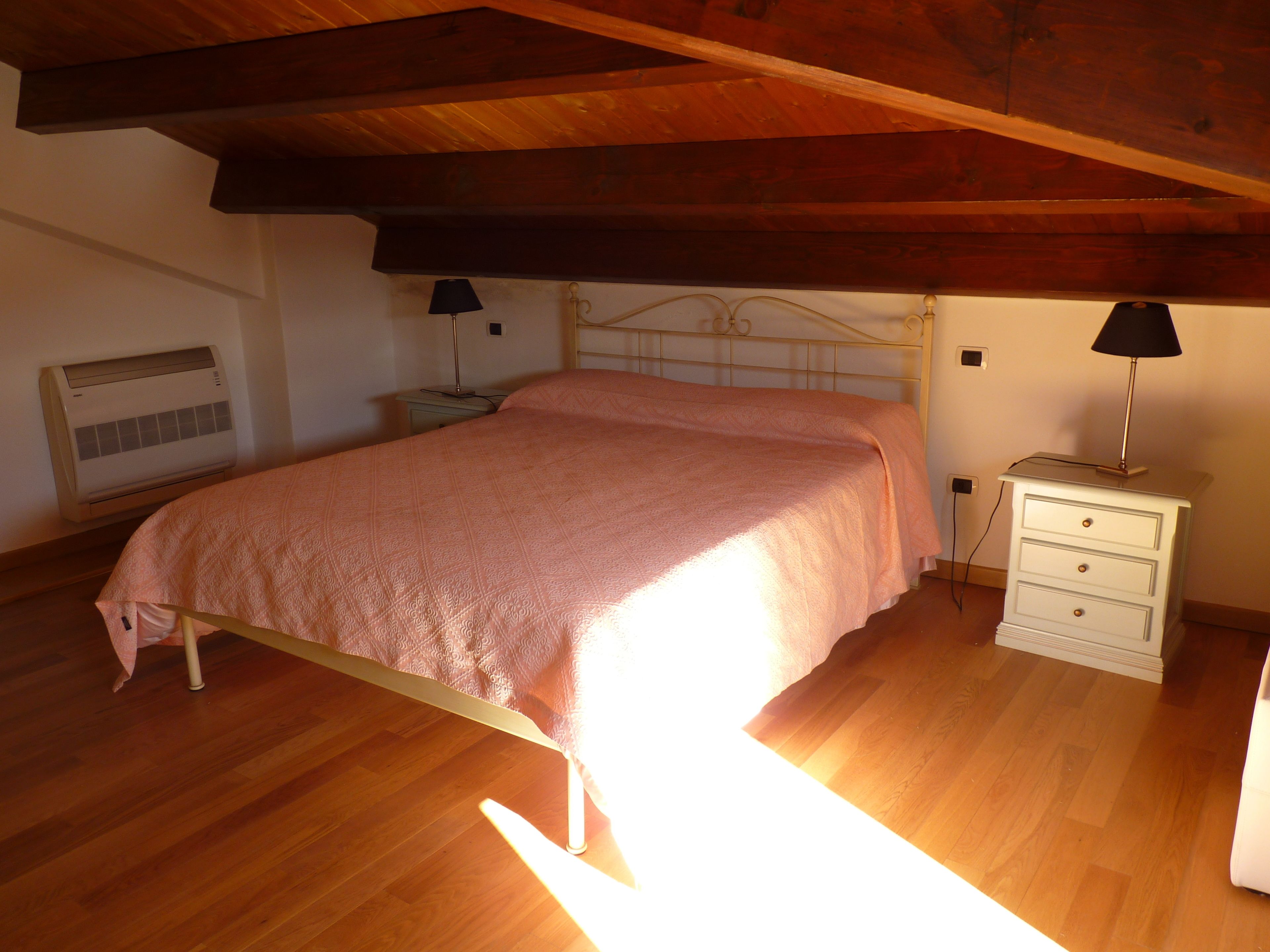 Ein Schlafzimmer mit Holzboden, einem Bett mit rosa Bettdecke, einem Nachttisch mit Lampe sowie einer Wand-Klimaanlage.