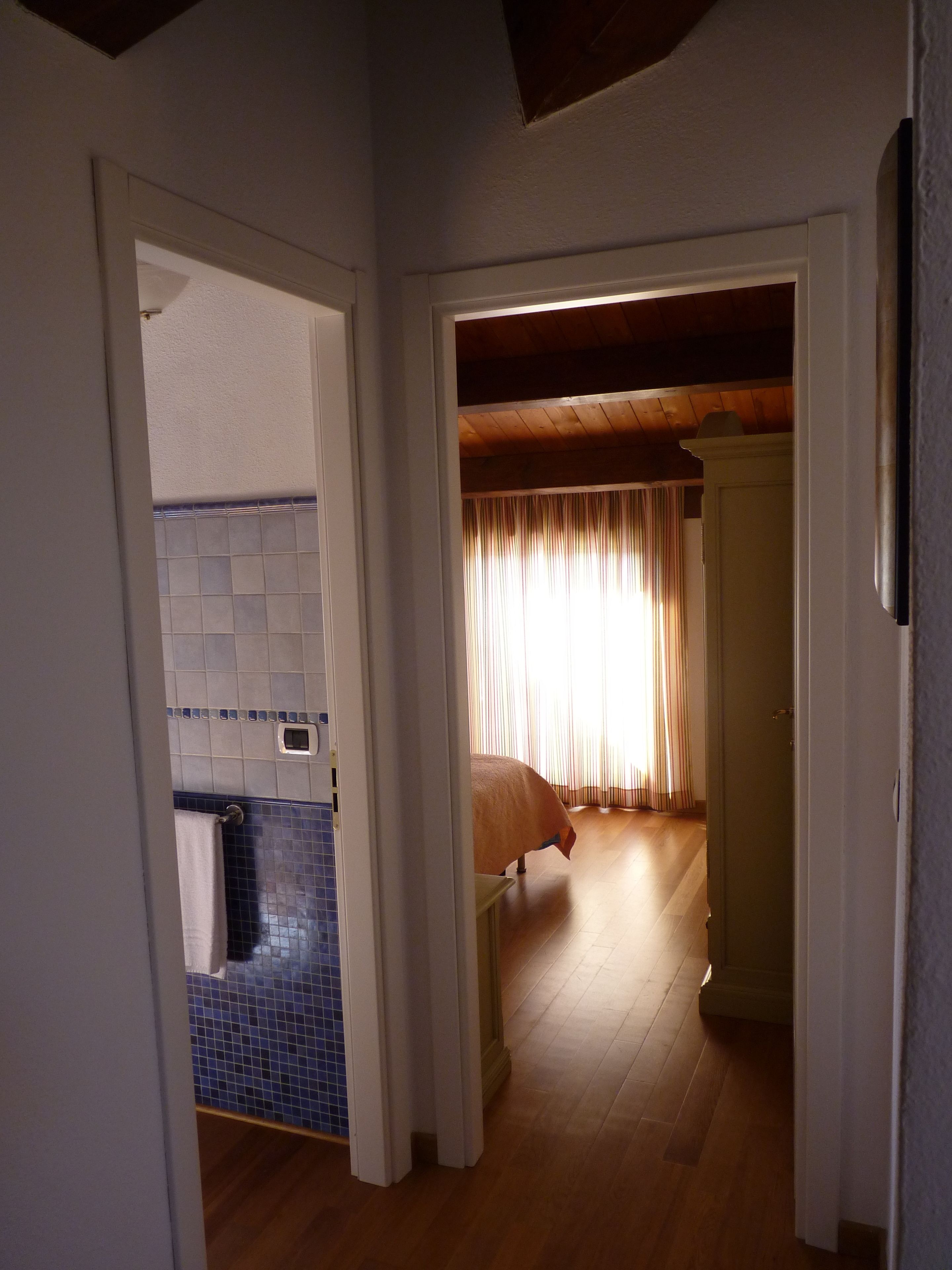 Un pasillo estrecho conduce a un dormitorio con cama y ventana con cortinas. A la izquierda se ve un baño con pared de azulejos y ducha.