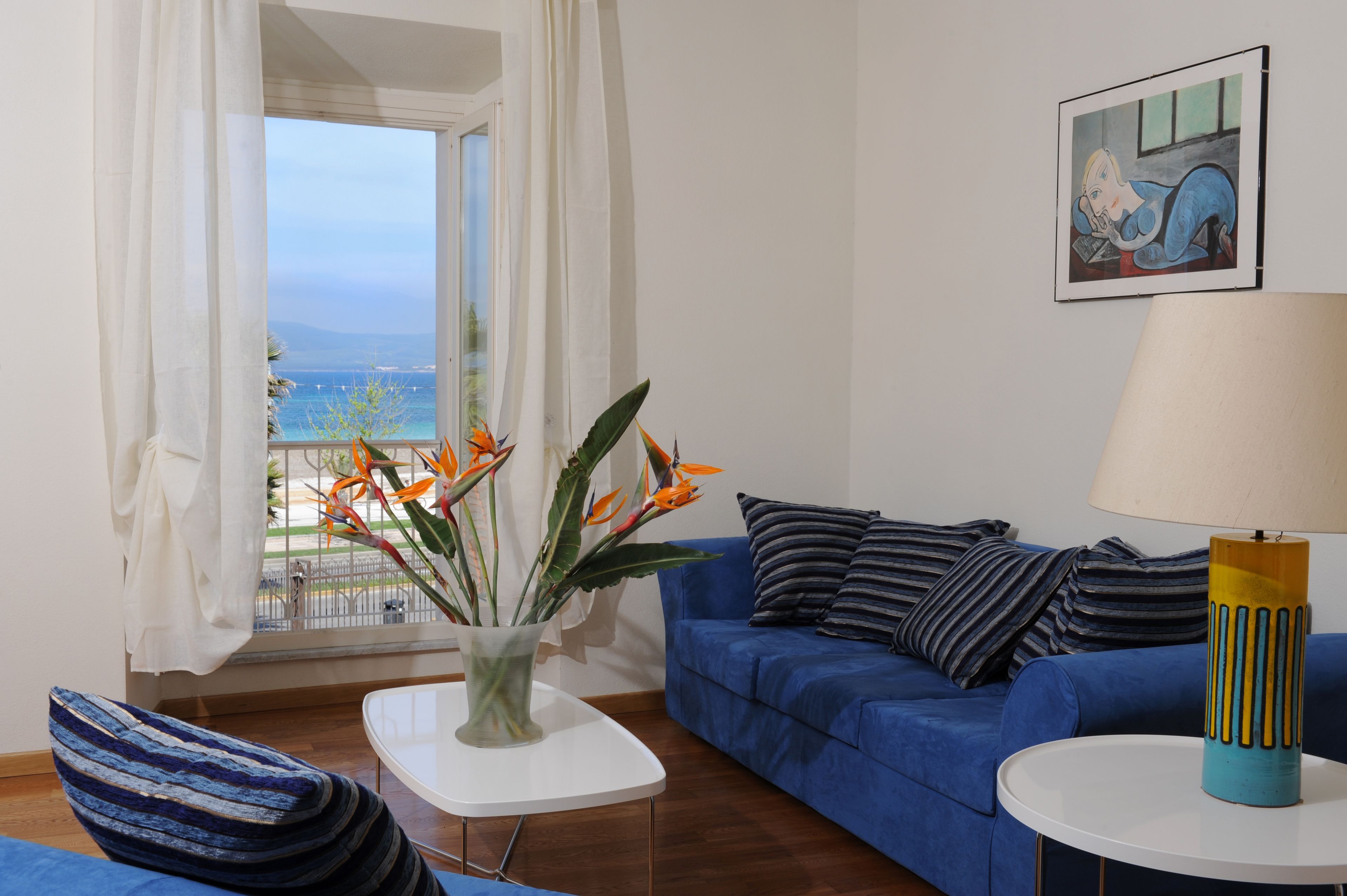 Un salon avec un canapé bleu, une table basse blanche, une lampe et une vue sur la mer par une fenêtre.