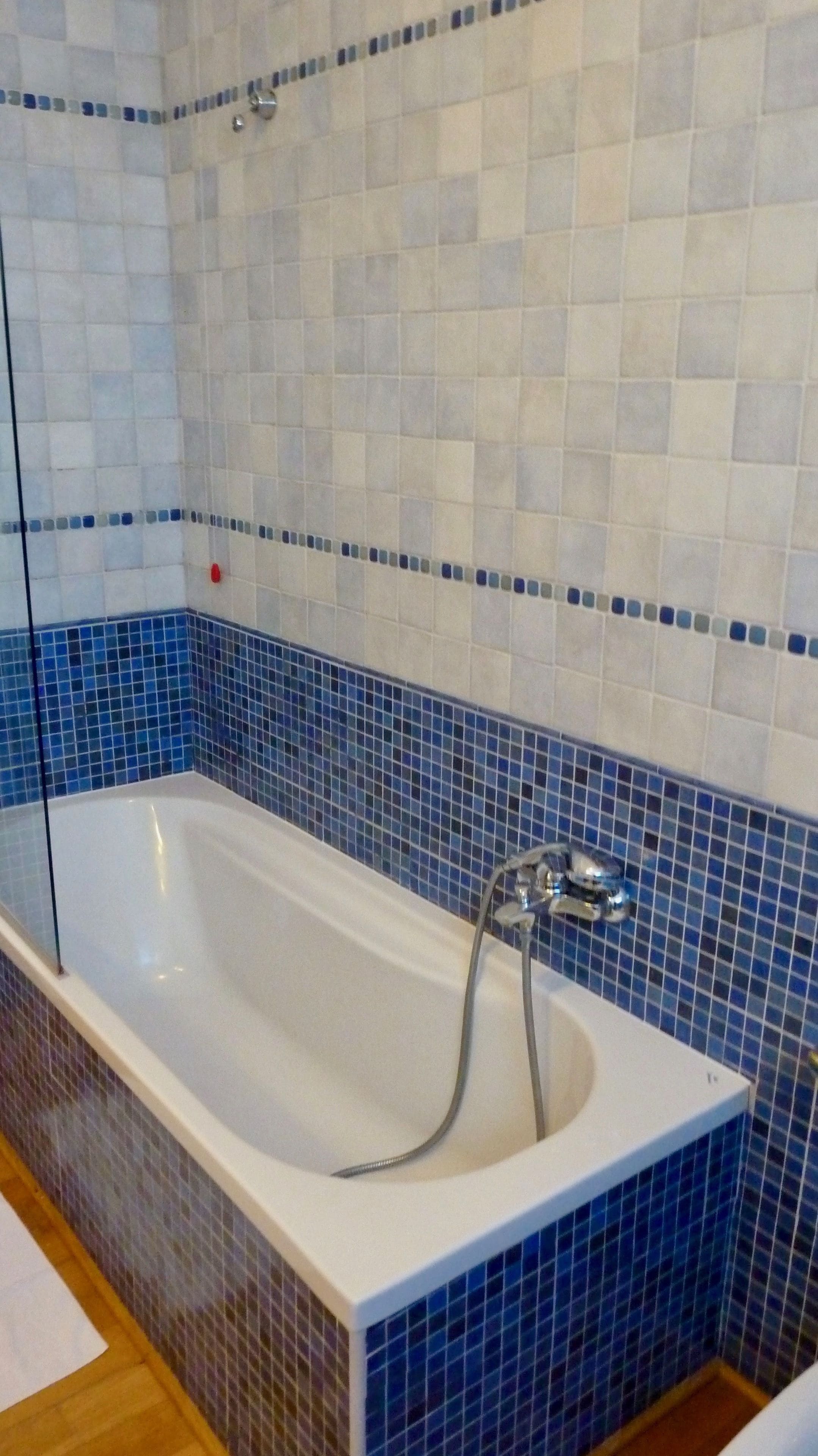 A bathroom with a white bathtub, blue mosaic tiles, and a wooden floor.