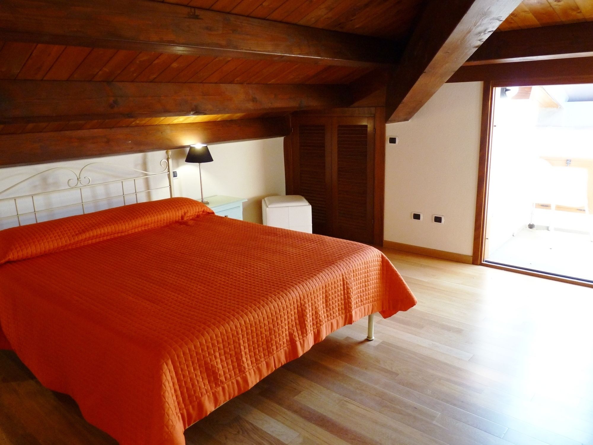 Ein Schlafzimmer mit Holzboden, einem Bett mit orangefarbener Bettdecke, einem Nachttisch, einer Lampe und einer Schiebetür, die auf einen Balkon führt.