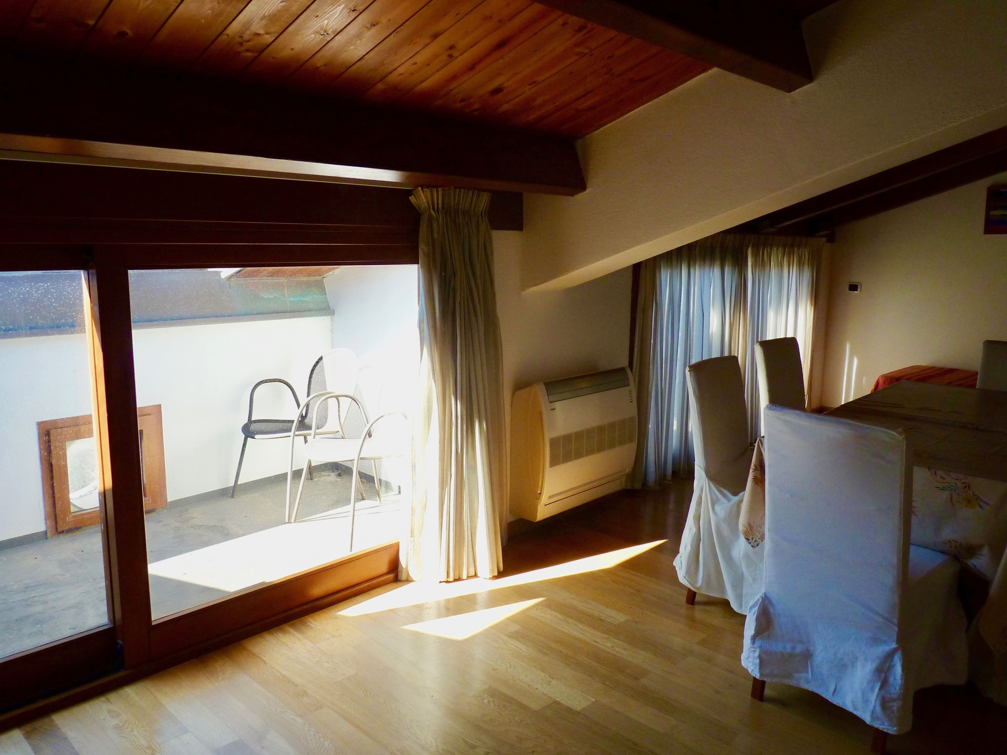 Une pièce avec un plafond en bois, un lit recouvert d’une courtepointe blanche, une chaise et une fenêtre avec des rideaux.
