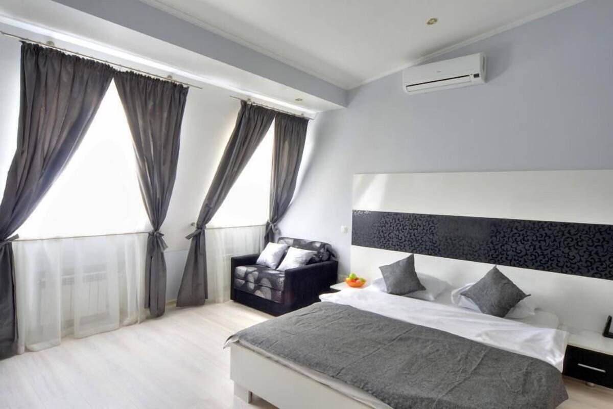 A modern bedroom with a large bed, a black sofa, and a window with sheer curtains.