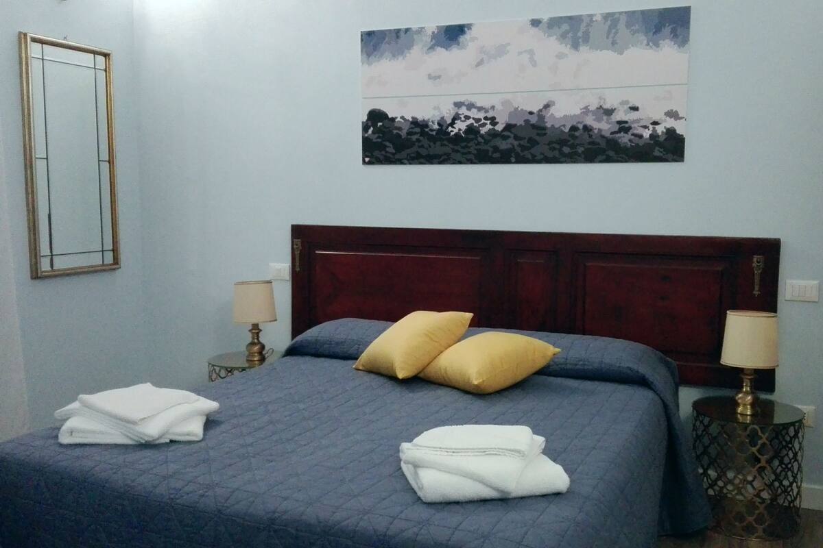 Ein ordentlich bezogenes Bett mit zwei Kissen, ein Nachttisch mit Lampe und ein Bild an der Wand.