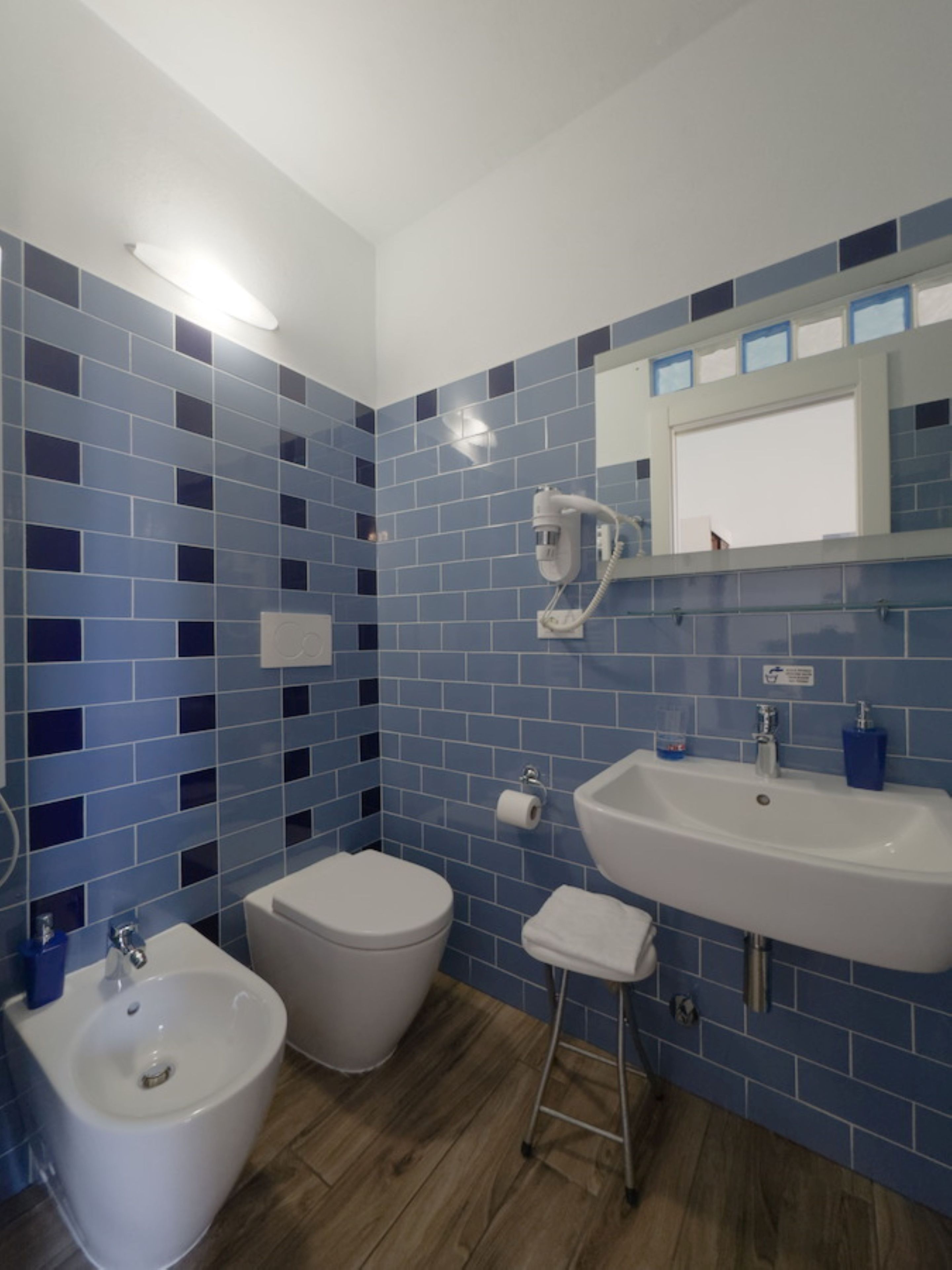 Un baño con bidé, inodoro y lavamanos, todo en blanco, con paredes de azulejos azules y suelo de madera.