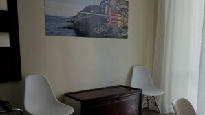 Une pièce comprenant un meuble en bois, deux chaises blanches, un tableau représentant une ville côtière et un luminaire noir fixé au mur.