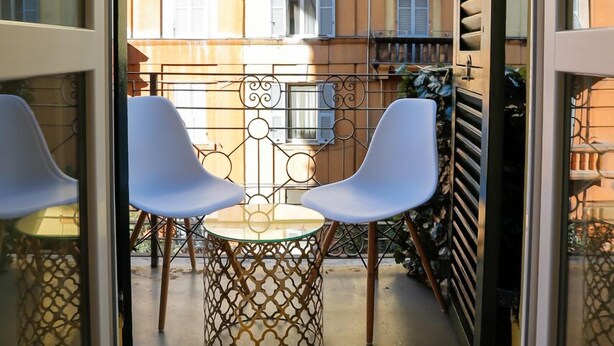 Un balcon avec deux chaises blanches et une petite table, donnant sur un quartier résidentiel où l’on aperçoit d’autres balcons et des fenêtres.