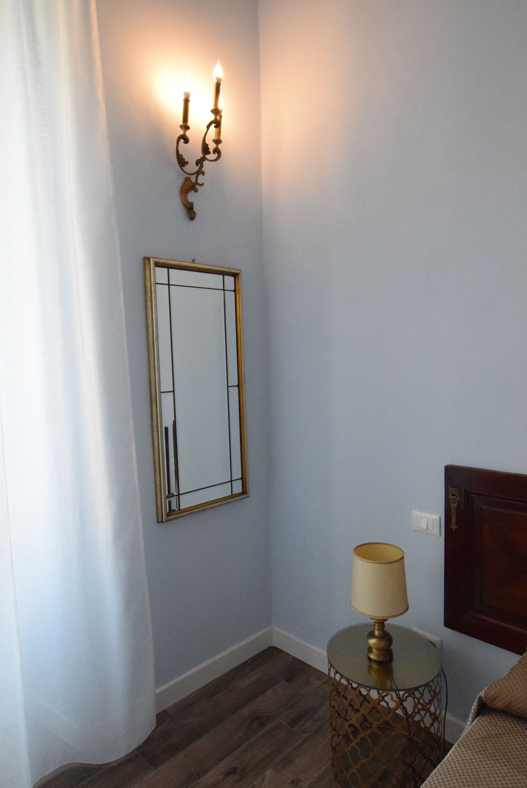 A room with a mirror, a wall-mounted light fixture, a round table with a lamp, and a wooden headboard.