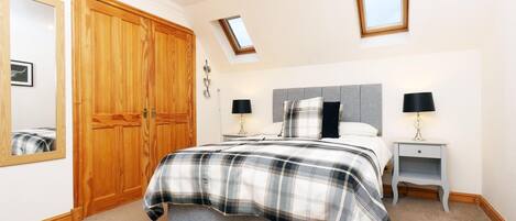 A bedroom with a large wooden wardrobe, a bed with a plaid duvet, two bedside tables with lamps, a mirror, and a skylight.