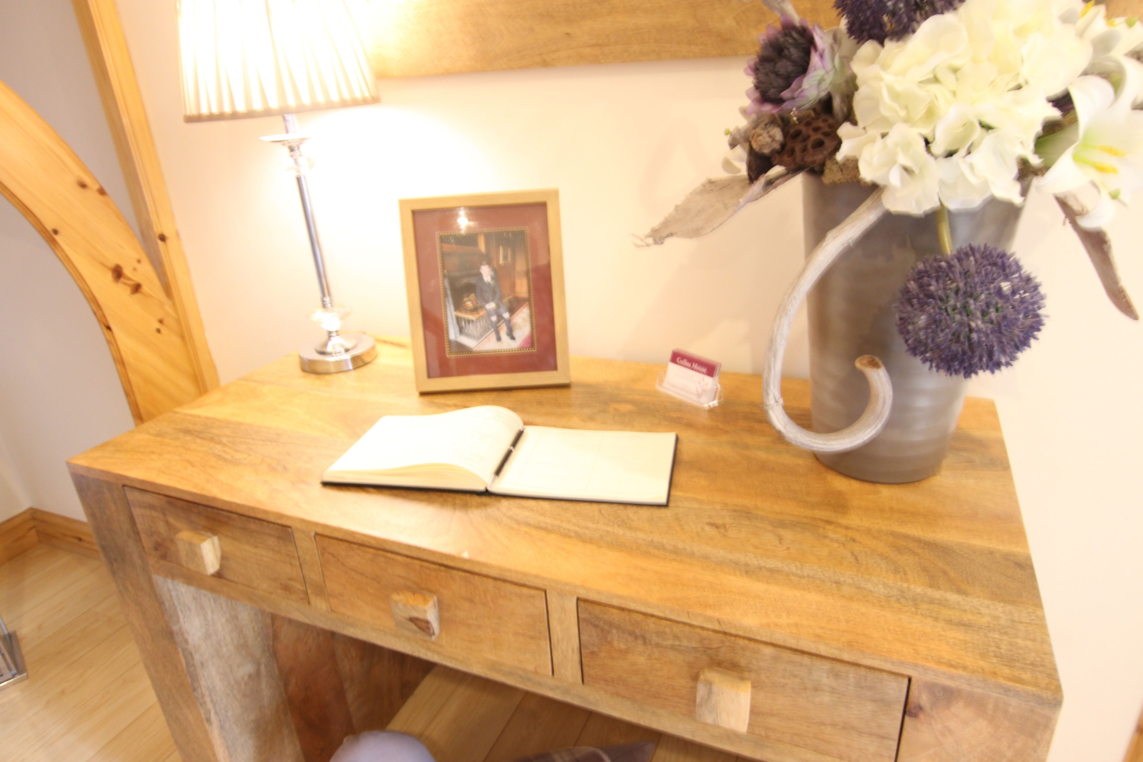 A wooden desk with a lamp, an open book, a framed picture, and a vase with flowers.