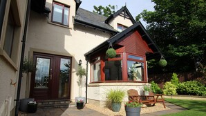 Une maison à deux étages avec un toit rouge, un porche à l’avant avec un banc et des plantes en pot.
