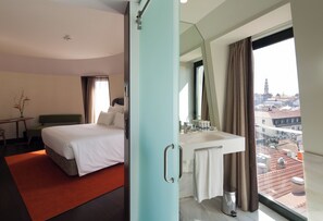 Une chambre d’hôtel avec un lit, une salle de bain dotée d’un lavabo et d’un miroir, et une vue sur la ville grâce à de grandes fenêtres.