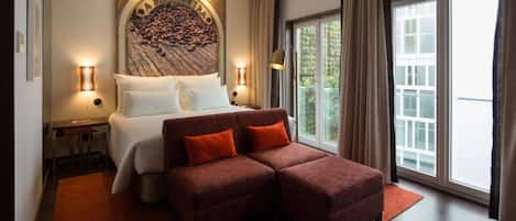 A hotel room with a bed, a sofa, a large window, and a wall-mounted artwork.