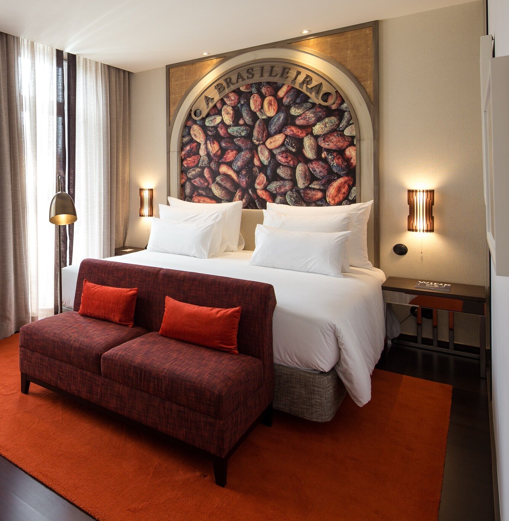 Una habitación de hotel con una cama, dos sofás rojos, un mural grande en la pared y dos lámparas de pared.