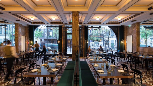 Een restaurant met een centrale kolom, meerdere tafels en uitzicht op de straat door grote ramen.