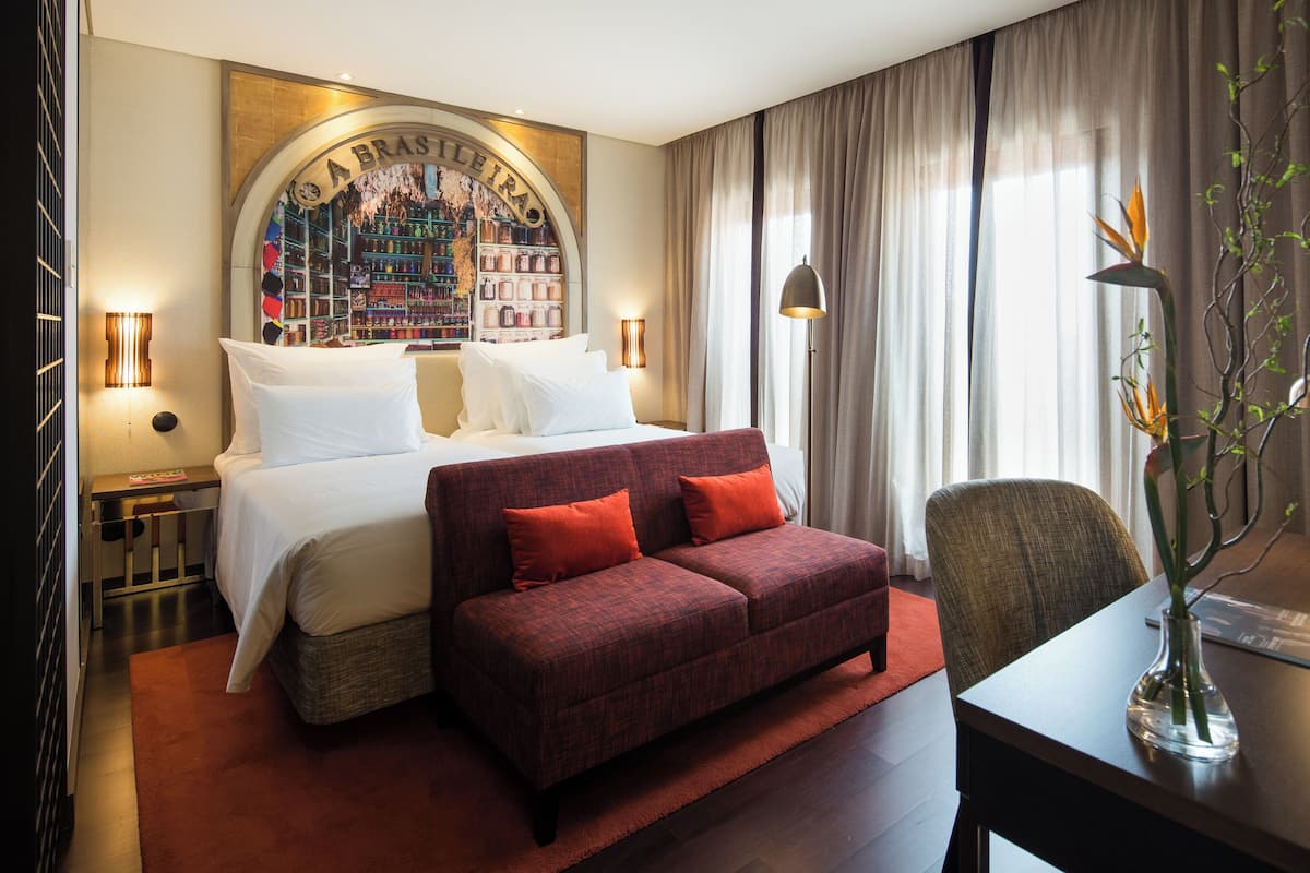 Habitación de hotel con cama, un sofá rojo, un escritorio con lámpara y un ventanal con cortinas.