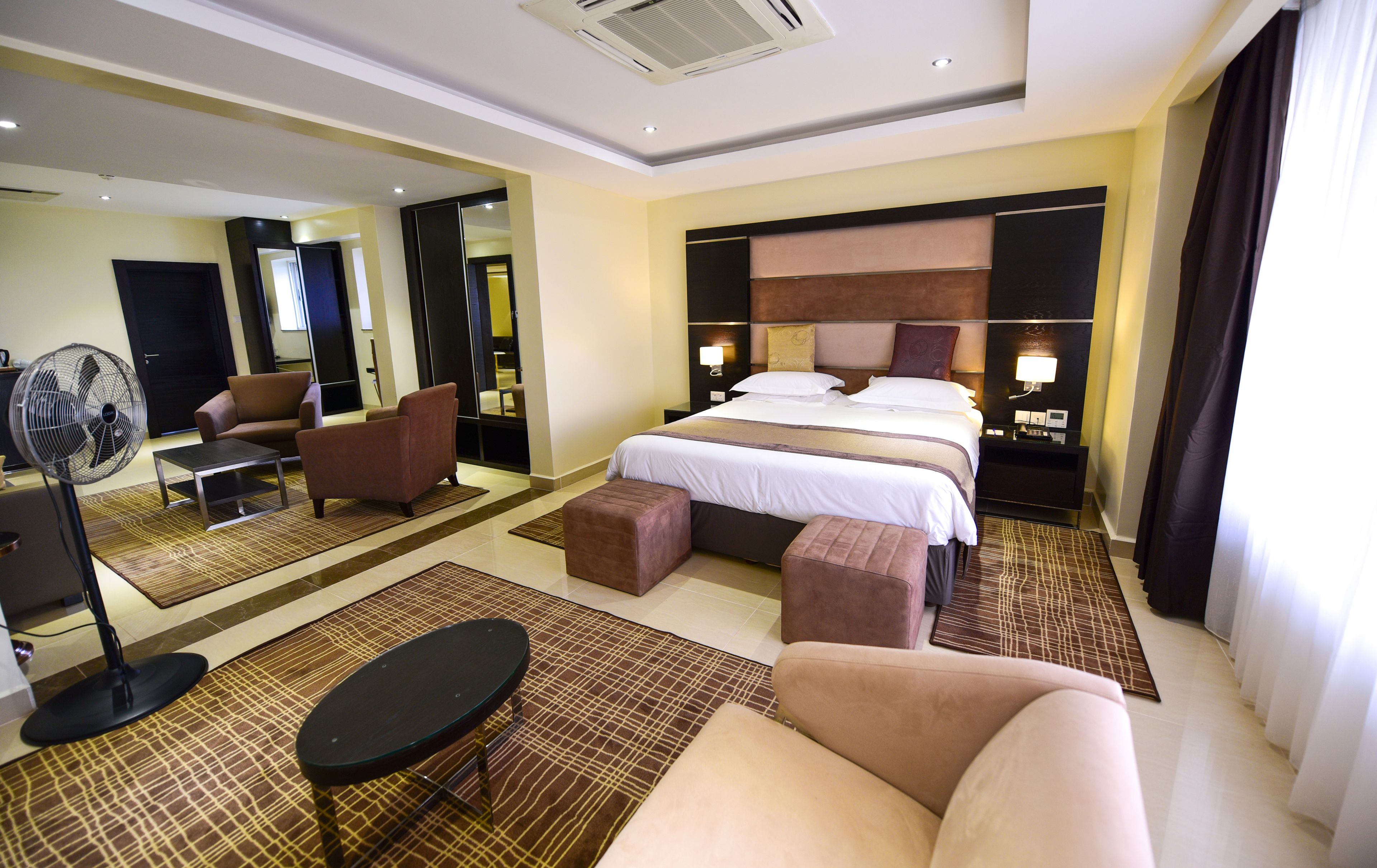 A hotel room with a bed, a sitting area with a sofa and armchair, a small table, a mirror, and a fan.