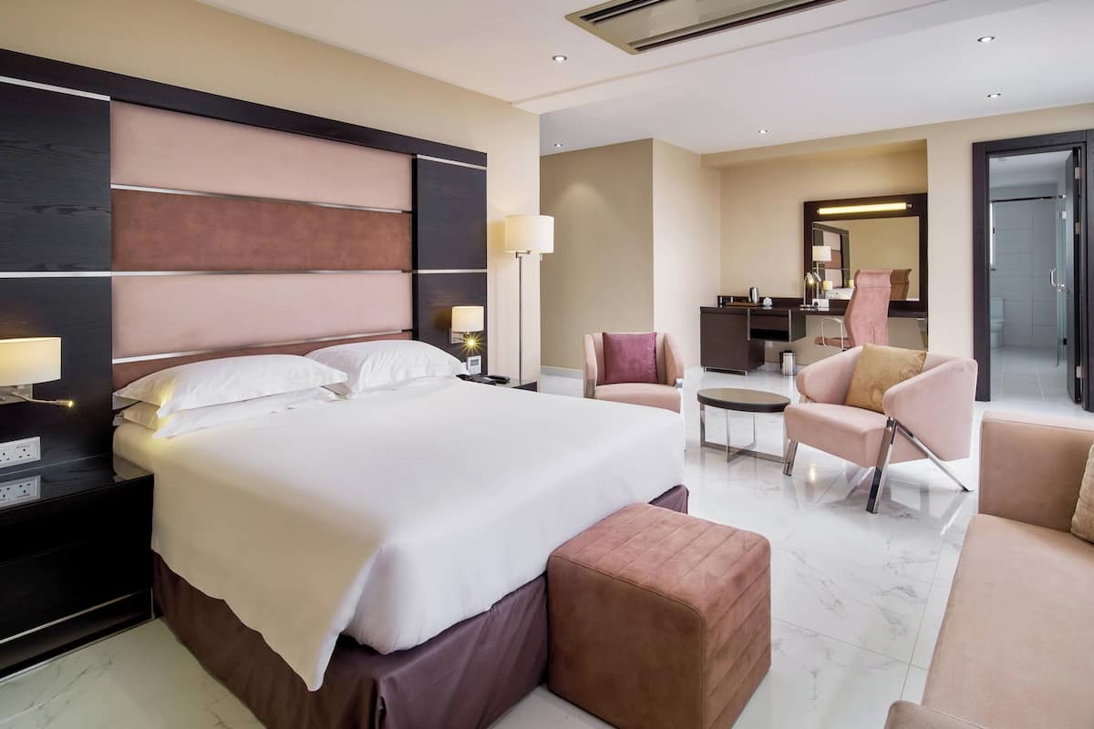 Une chambre d’hôtel moderne dotée d’un grand lit, d’un coin salon avec un canapé et un fauteuil, d’une petite table et d’une salle de bain séparée avec un miroir et un lavabo.