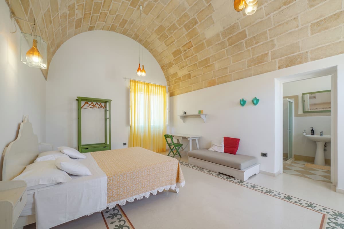 Une chambre à coucher avec un lit, une chaise, un canapé, un miroir et une salle de bain visible à travers une porte ouverte.