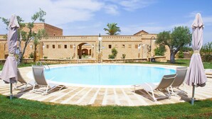 Une piscine entourée de chaises longues et de parasols, bordée d’un bâtiment en pierre doté d’arcades, sous un ciel dégagé.