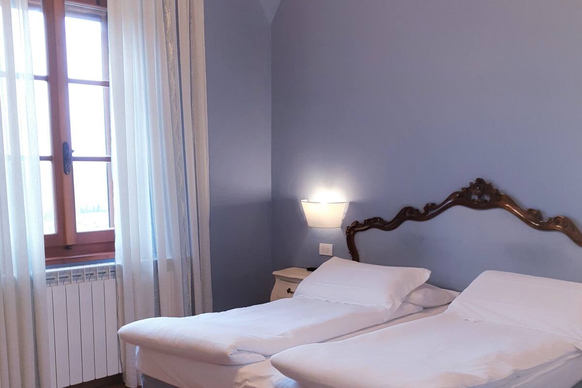 A bedroom with a bed, a nightstand, a wall-mounted lamp, a window with curtains, and a radiator.
