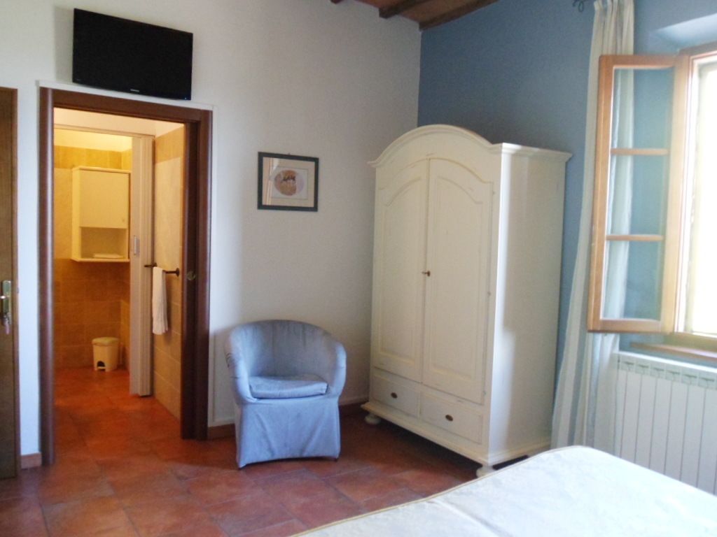 A bedroom with a bed, a white wardrobe, a blue armchair, and a door leading to a bathroom.