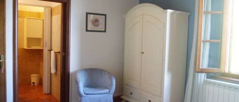 A bedroom with a bed, a white wardrobe, a blue armchair, and a door leading to a bathroom.